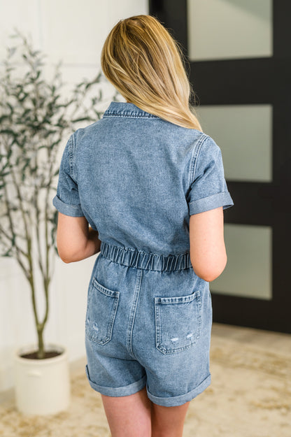 Person wearing a denim romper standing in a room with a plant and framed picture.