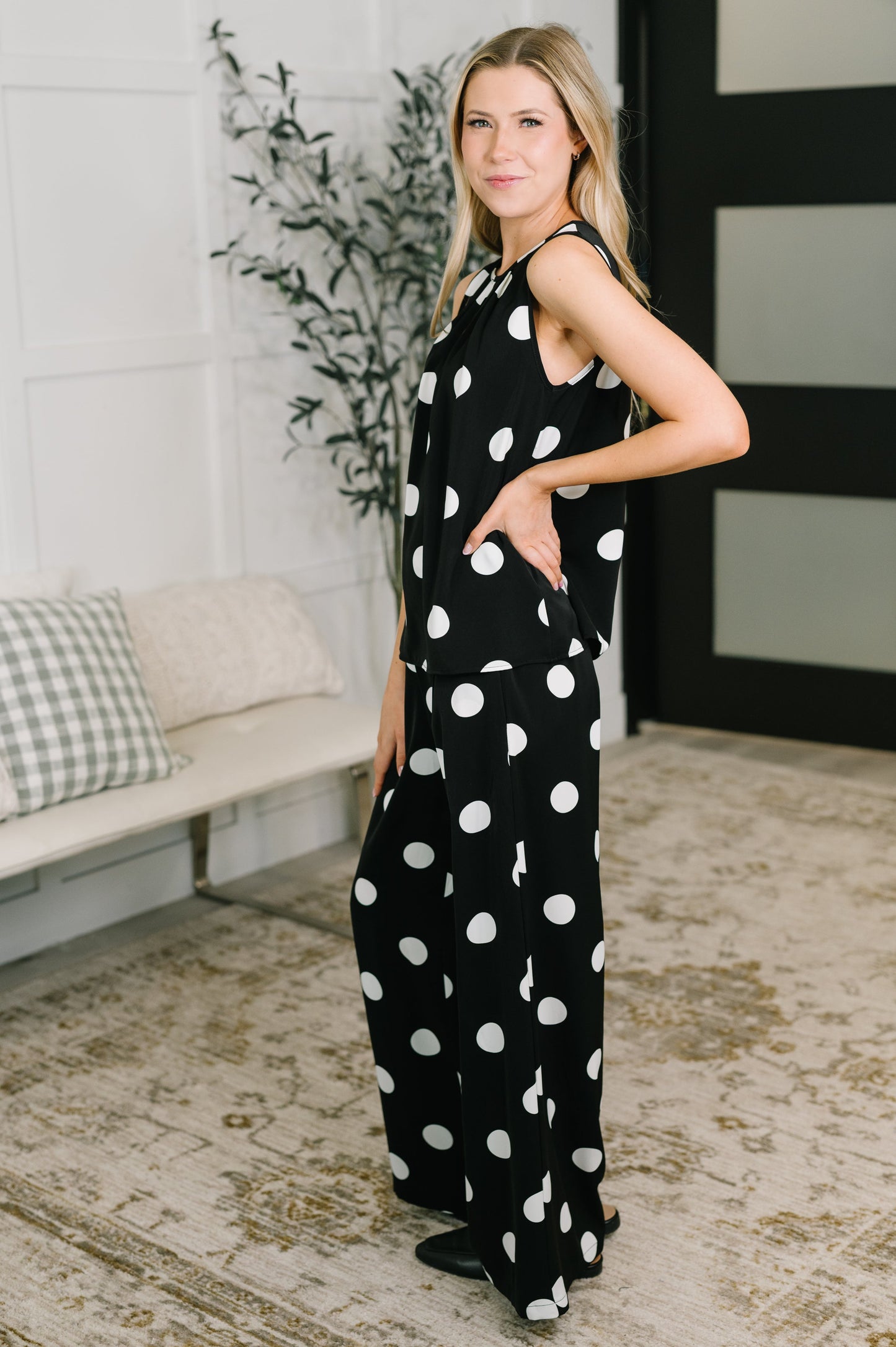 Woman wearing a black polka dot jumpsuit standing in a room with a couch and plant.
