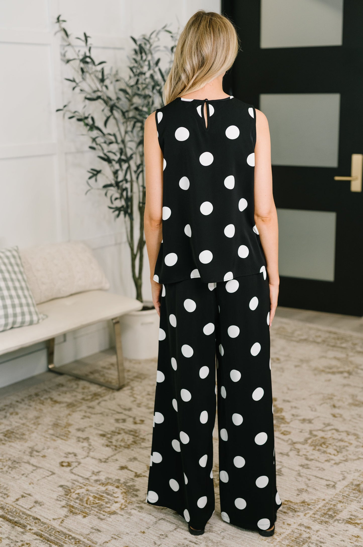 Person wearing a black polka dot jumpsuit standing in a room with a plant and bench.