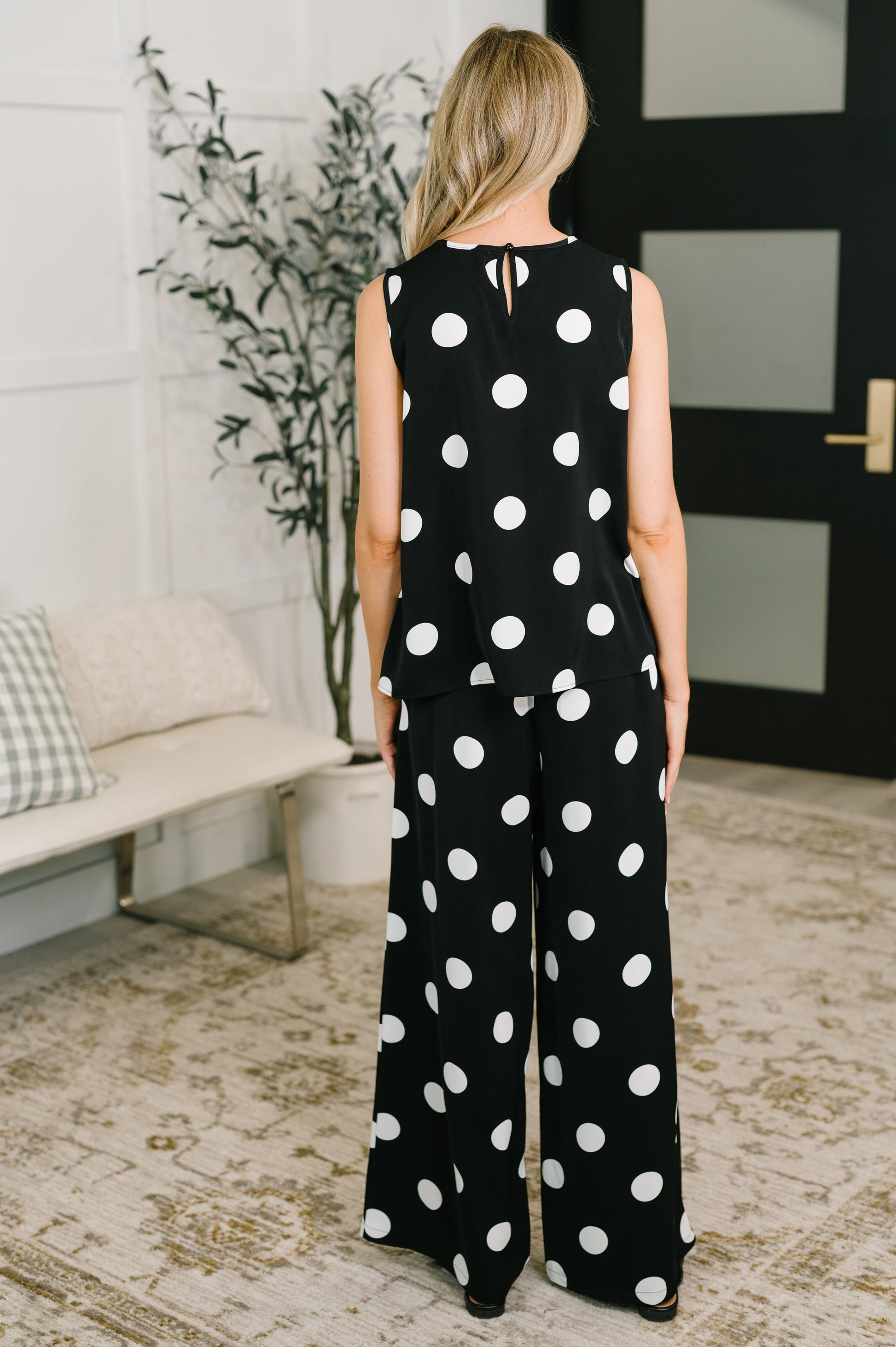 Person wearing a black polka dot jumpsuit standing in a room with a plant and bench.