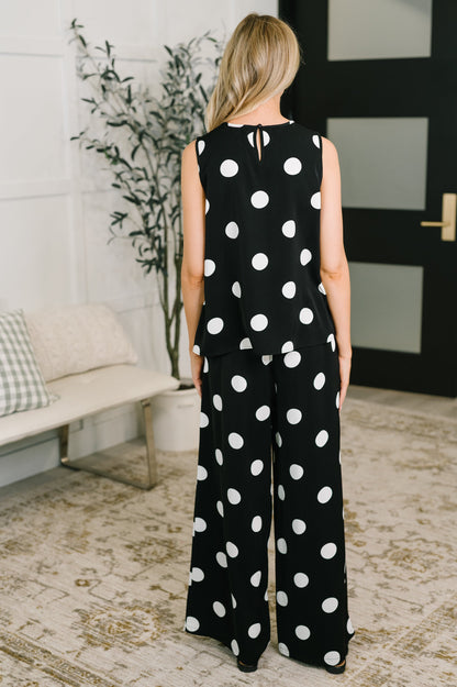 Person wearing a black polka dot jumpsuit standing in a room with a plant and bench.