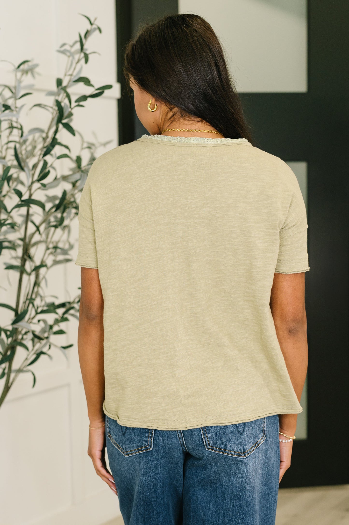 Woman wearing a beige top and blue jeans standing indoors with a plant in the background