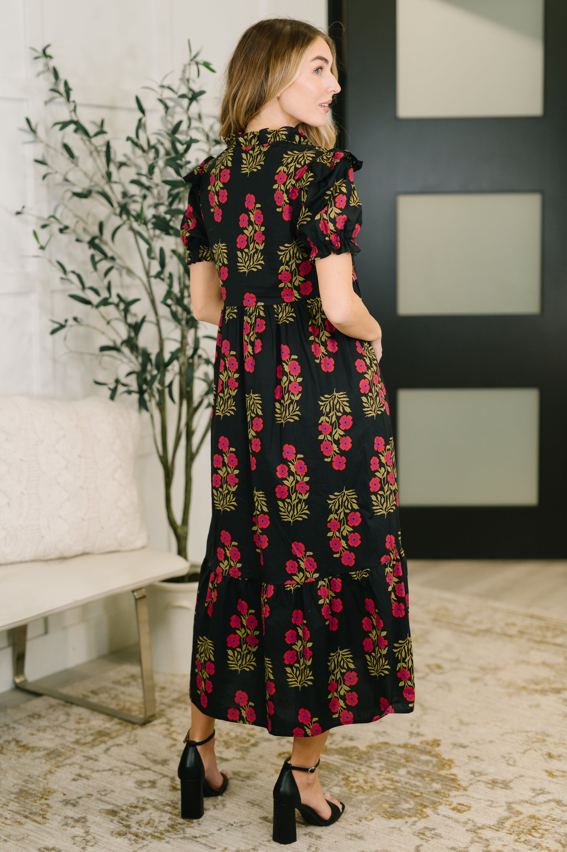 Woman wearing a black dress with red floral pattern in a room with a plant and sofa.