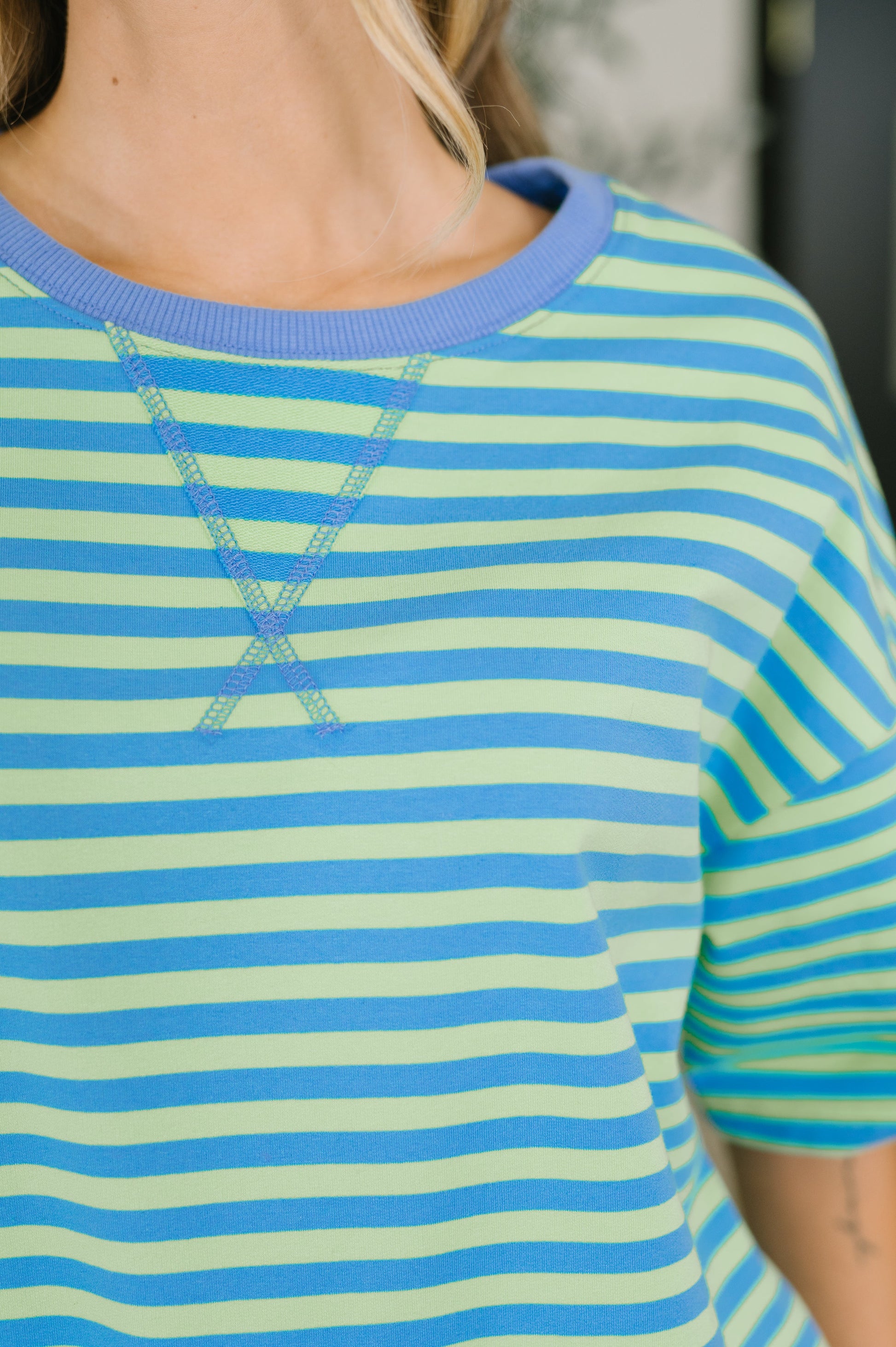 Close-up of a person wearing a short-sleeve crewneck top with horizontal light green and medium blue stripes, plus a light blue ribbed collar.