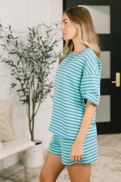 A woman wearing a matching two-piece lounge set with light blue and white horizontal stripes, standing in a brightly lit room with a potted plant and a bench in the background.