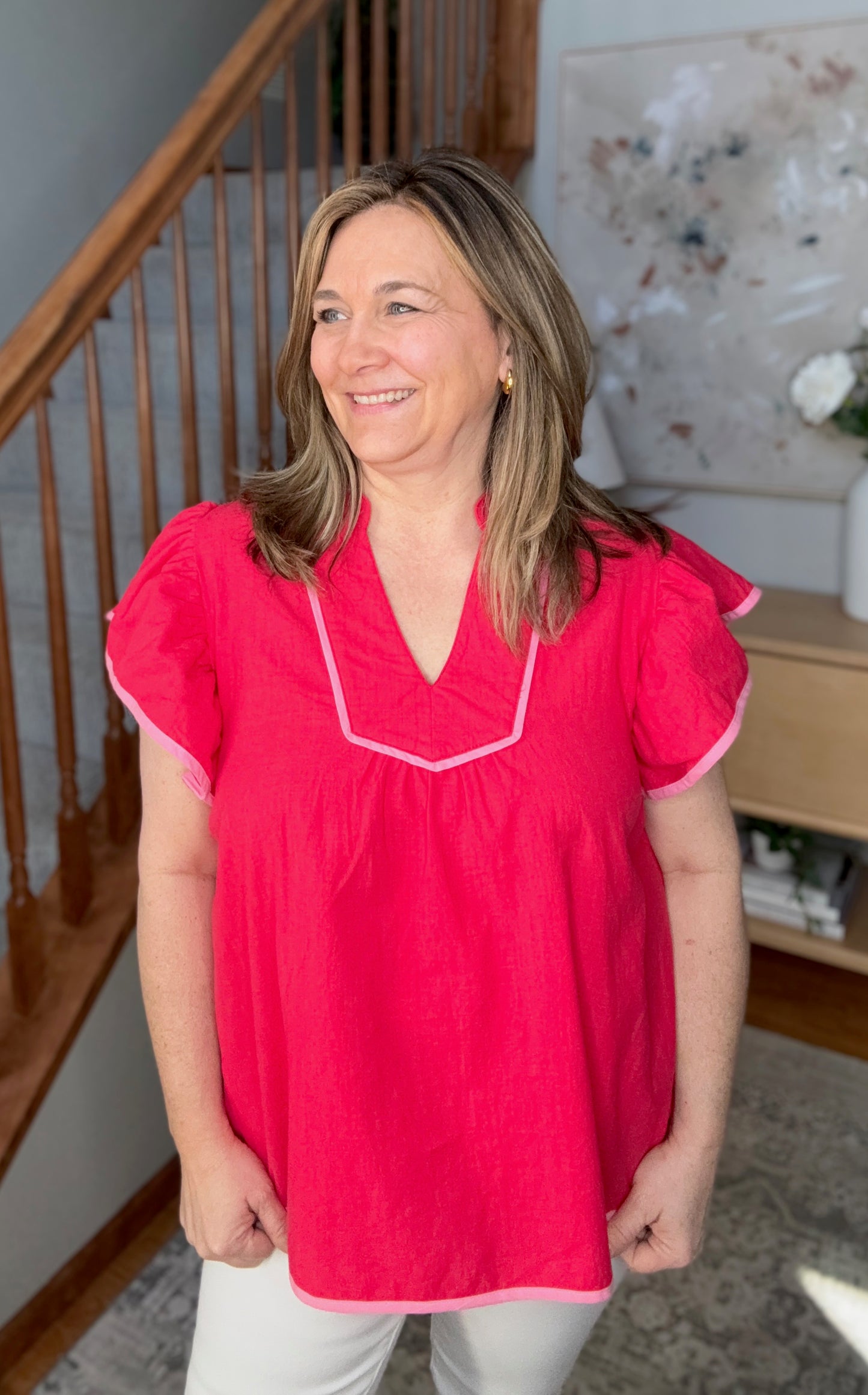 Woman wearing a pink blouse standing in a home setting with a staircase and decor in the background.