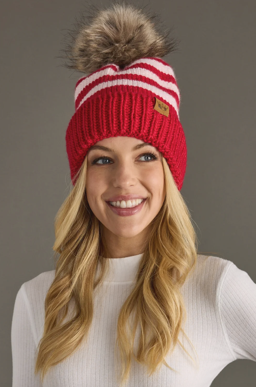 Woman wearing a red and white striped knit beanie with a fur pom-pom against a gray background