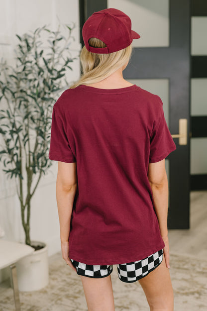 Person wearing a maroon t-shirt and checkered shorts, standing in a room with a plant and door.
