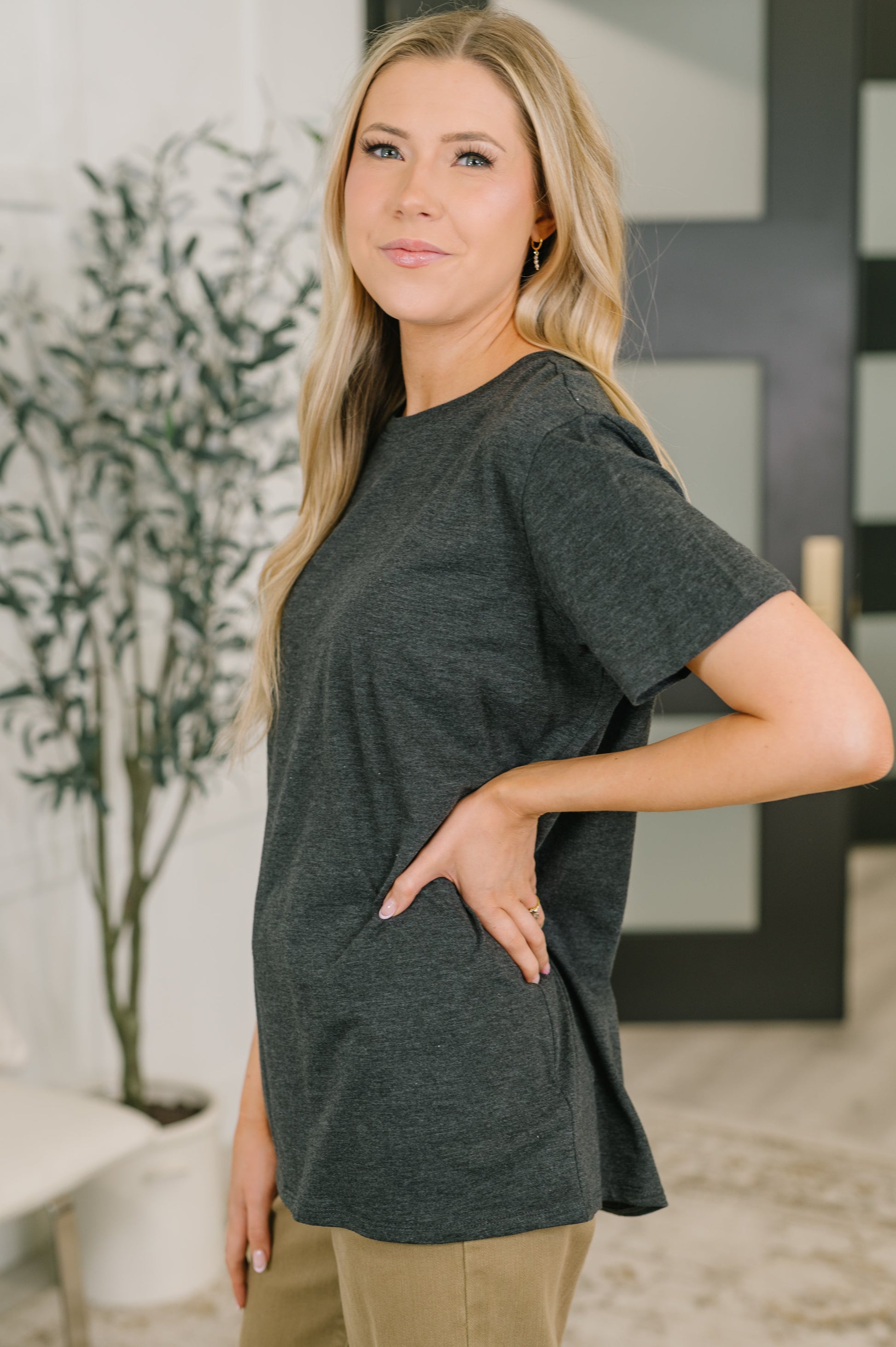 Woman wearing a dark gray t-shirt standing indoors with a plant and wall art in the background.
