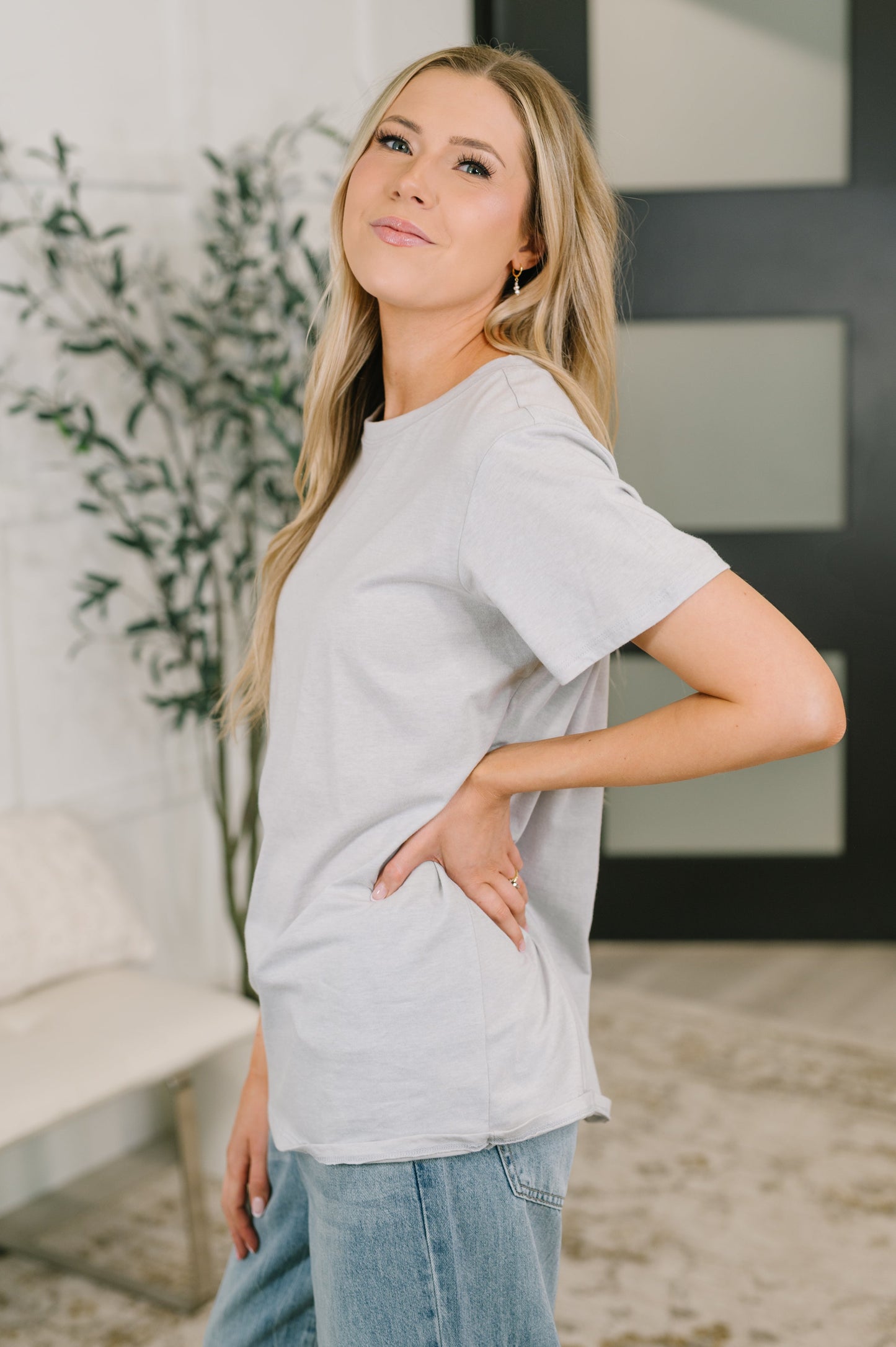 Woman wearing a light gray t-shirt and blue jeans standing indoors with a plant in the background.