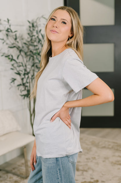 Woman wearing a light gray t-shirt and blue jeans standing indoors with a plant in the background.