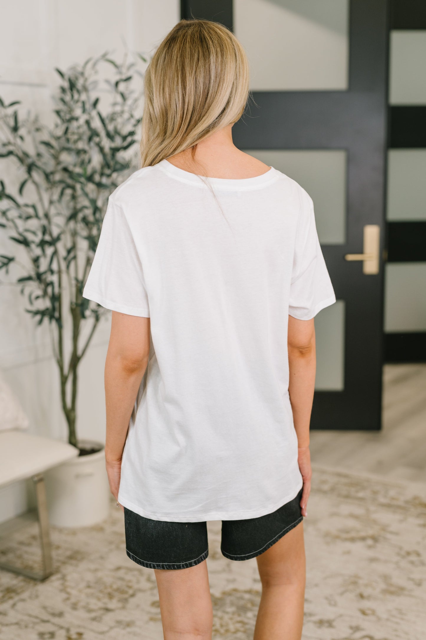 Person wearing a white t-shirt and black shorts standing in a room with a plant and door in the background.