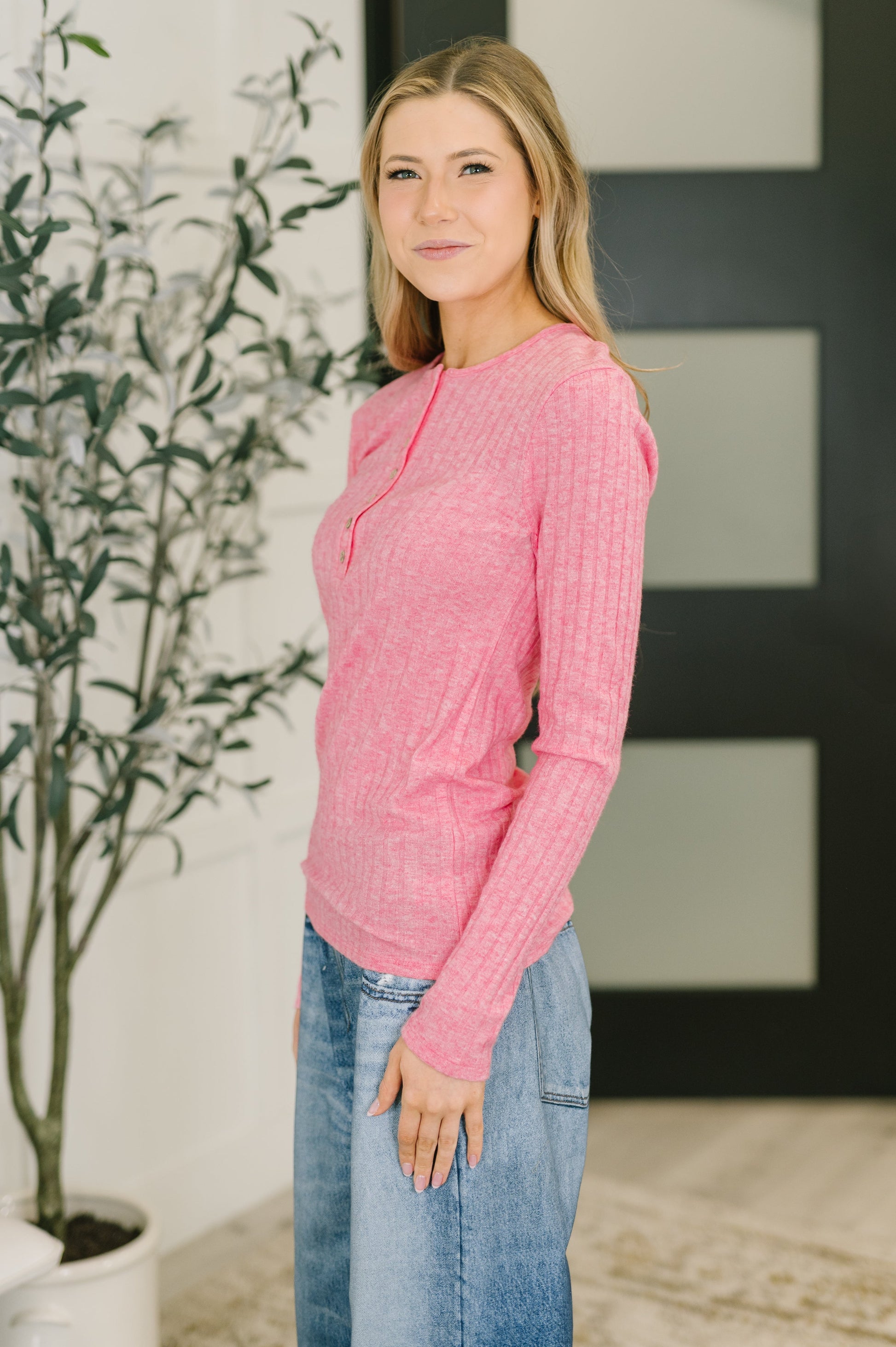 Woman wearing a pink sweater and blue jeans standing indoors with a plant in the background