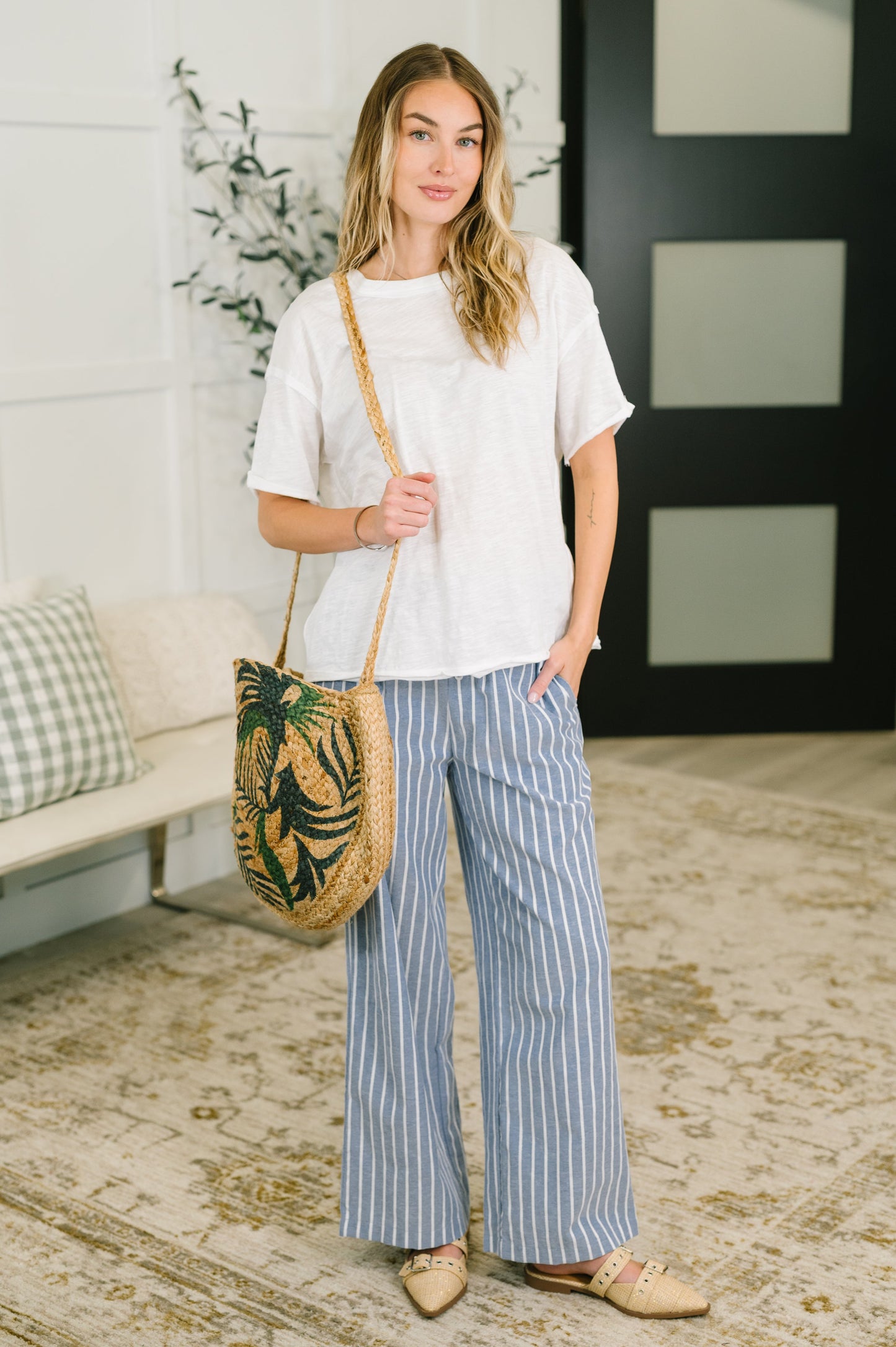 Woman holding a woven bag with a plant design in a room with a white wall and decorative elements.