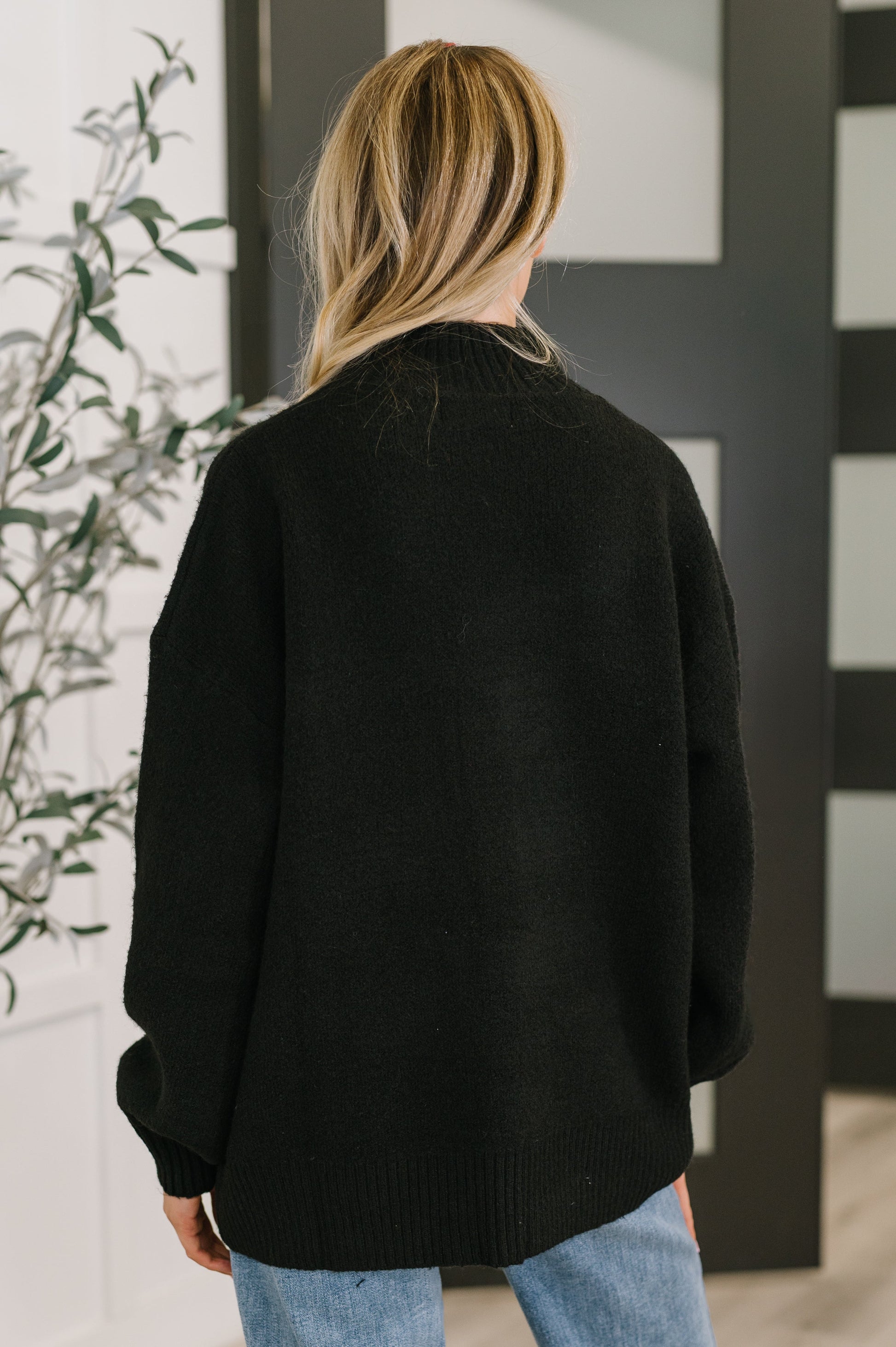 Person wearing a black sweater in an indoor setting with a plant and striped wall.