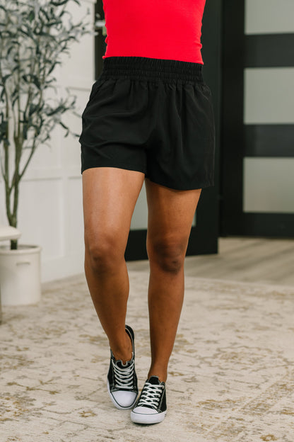 Person wearing black shorts and black sneakers with white soles on a stone pavement.