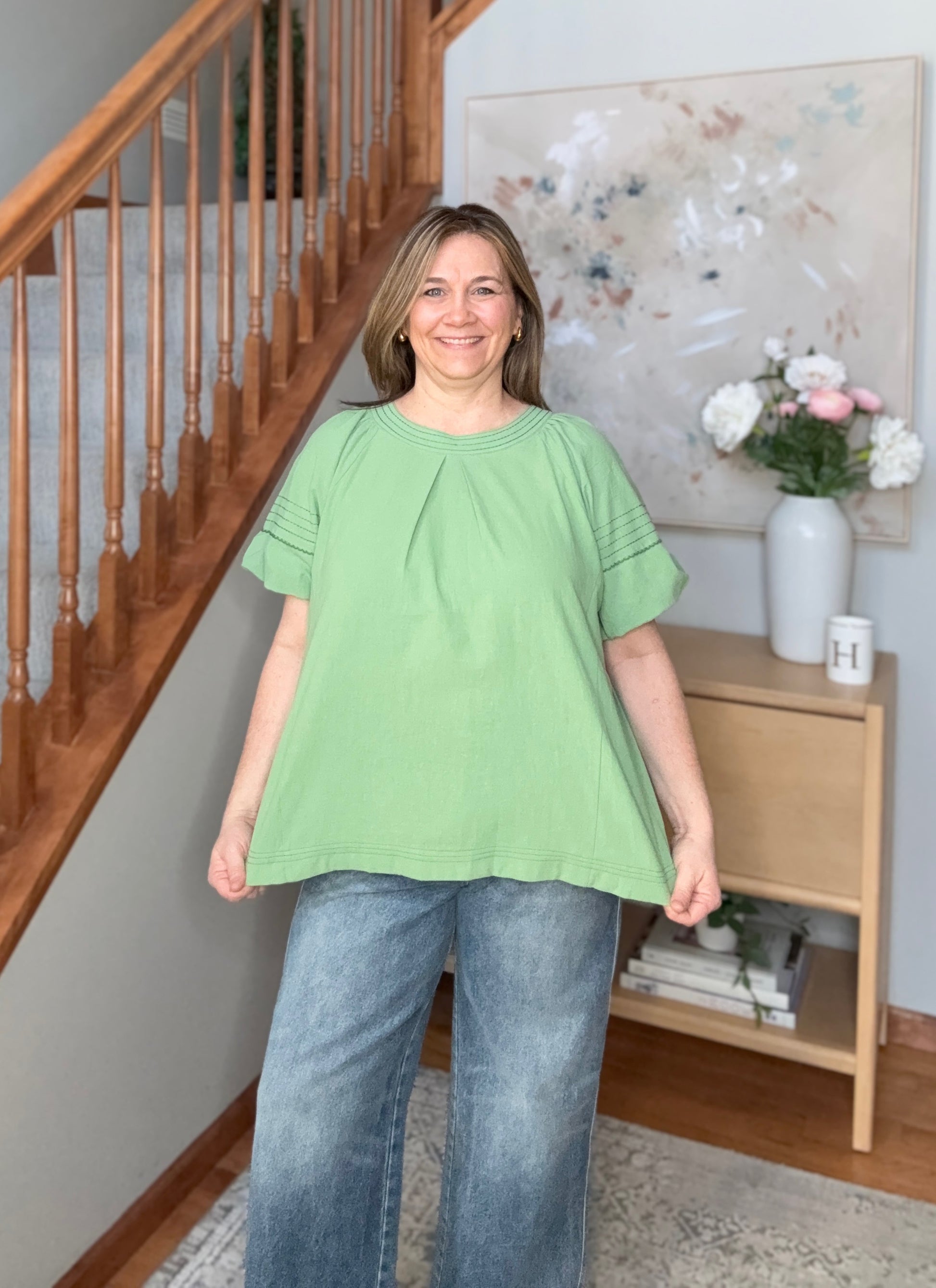 Woman wearing a green blouse and blue jeans standing in a home setting with a staircase and decor.