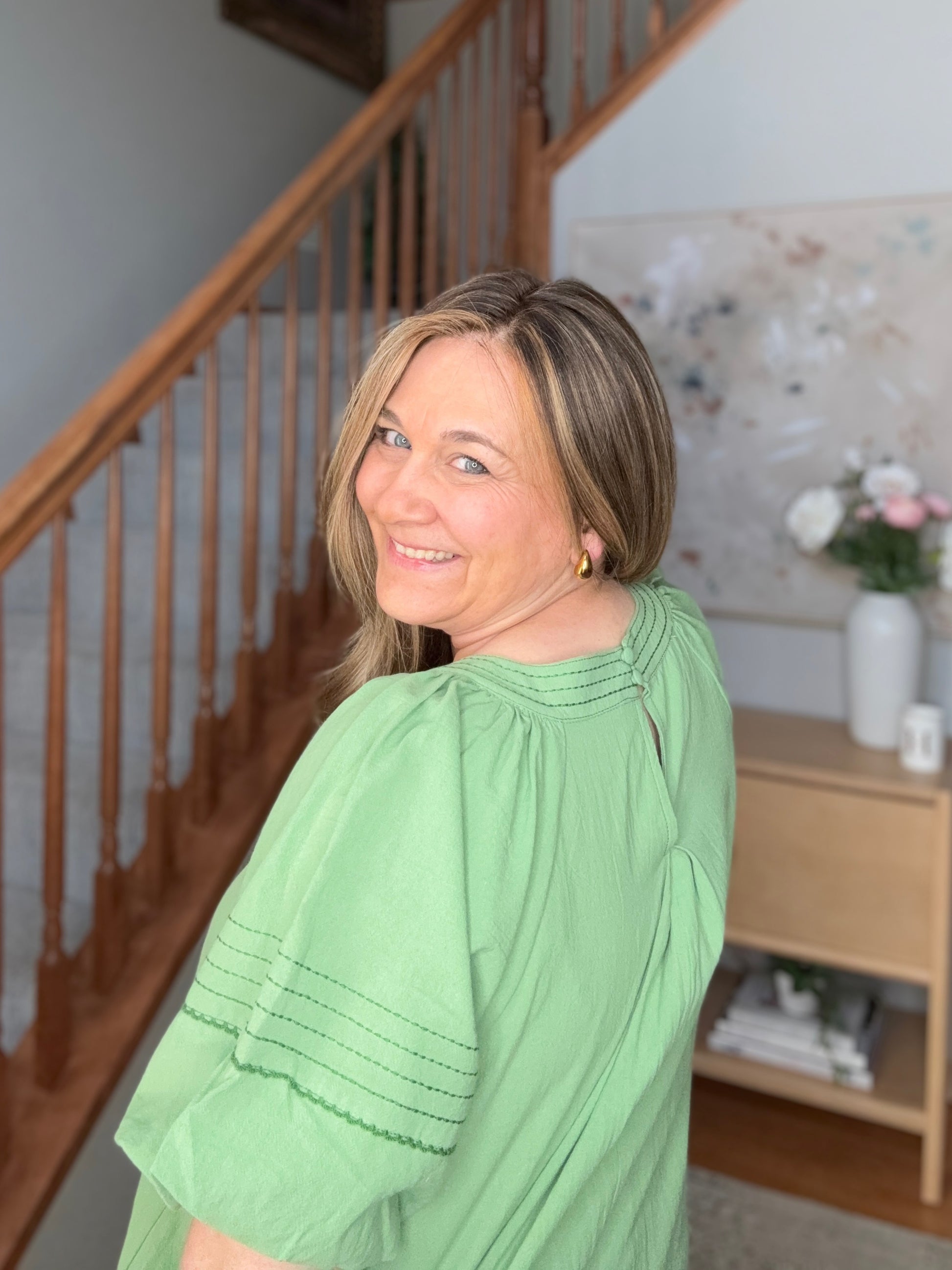 Woman in a green dress standing in a home interior with wooden staircase and decor.