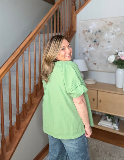Woman in a green shirt standing in a home interior with wooden staircase and decor.