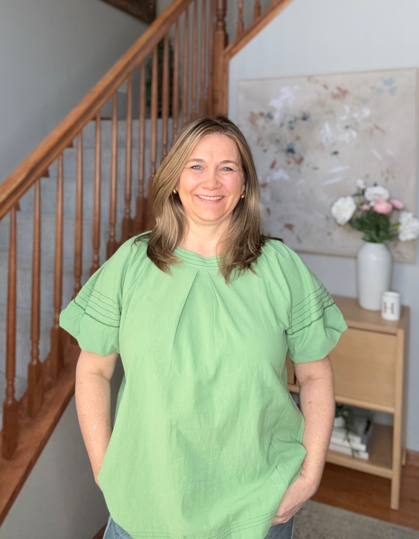 Woman wearing a green shirt standing in a home interior with a staircase and decorative items.