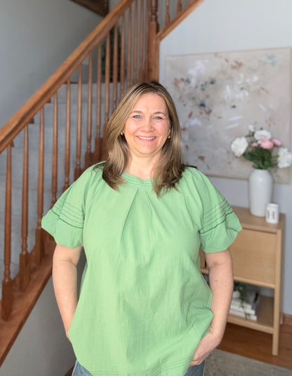 Woman wearing a green shirt standing in a home interior with a staircase and decorative items.