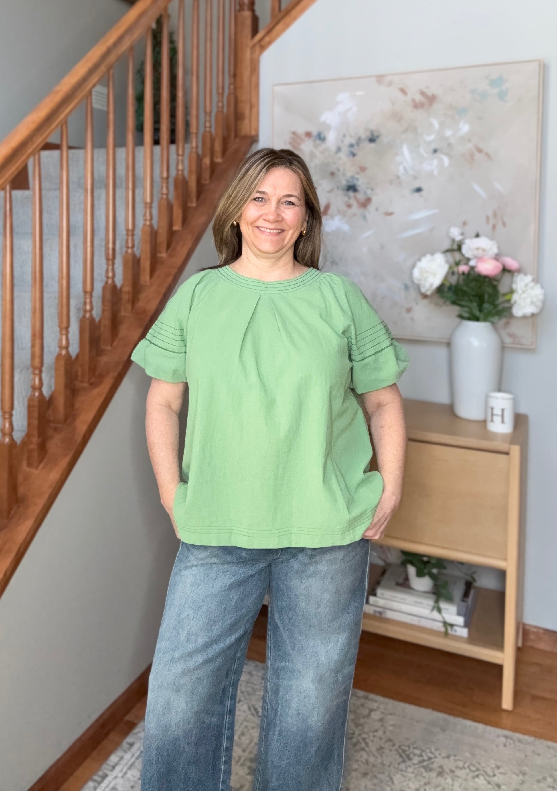 Woman in a green top and blue jeans standing in a home setting with a staircase and decor.