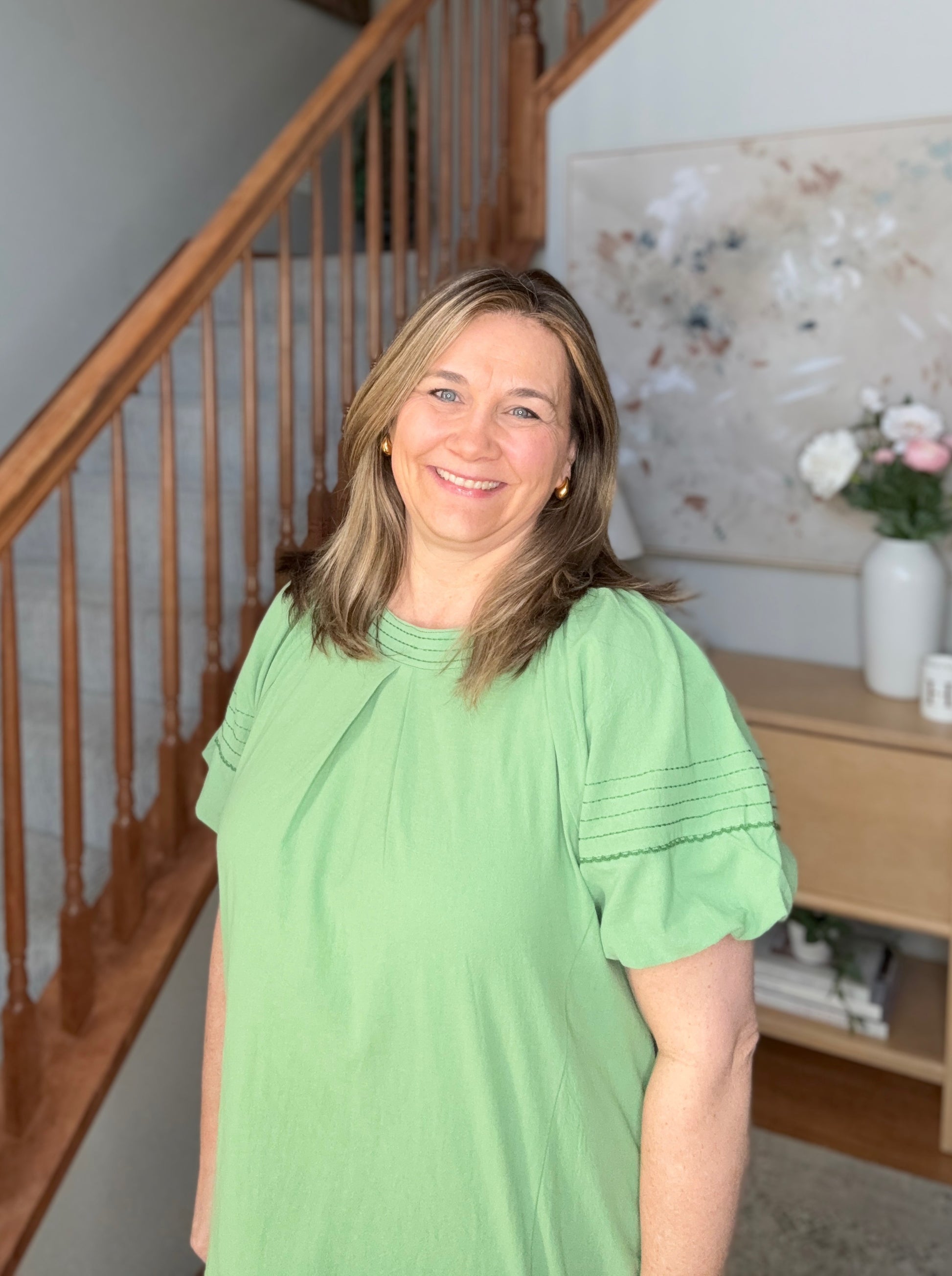 Woman in a green shirt standing in a home interior with a staircase and decorative items.