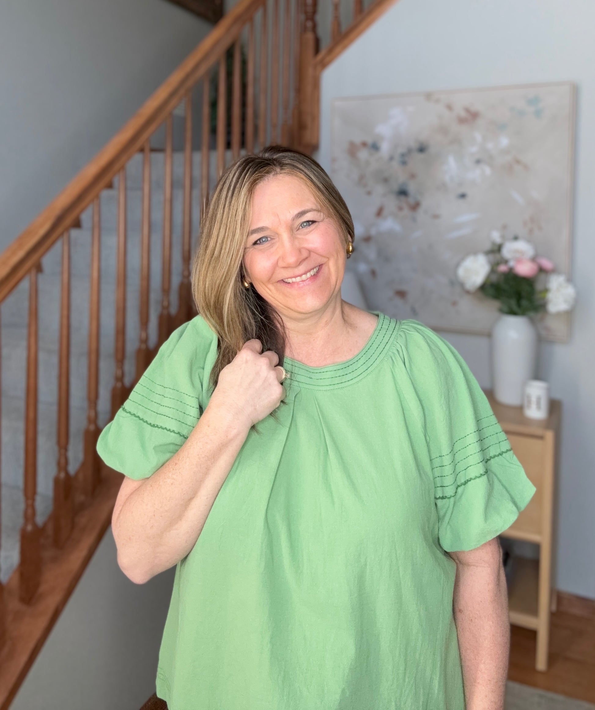 Woman in a green dress standing in a home setting with a staircase and decor.