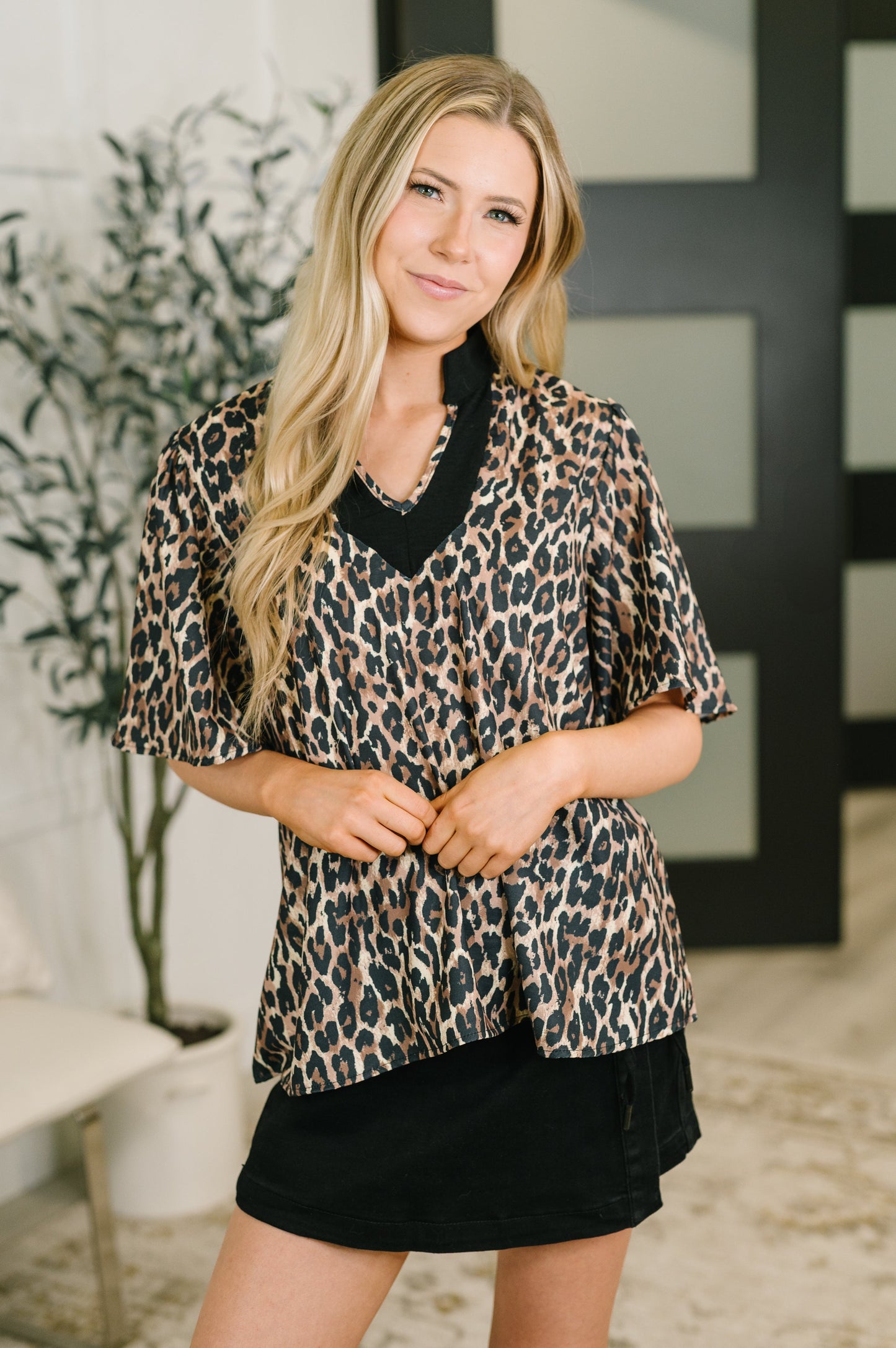 A woman modeling a leopard print short-sleeve blouse with a black V-neck trim, paired with a black skirt, in an indoor setting.