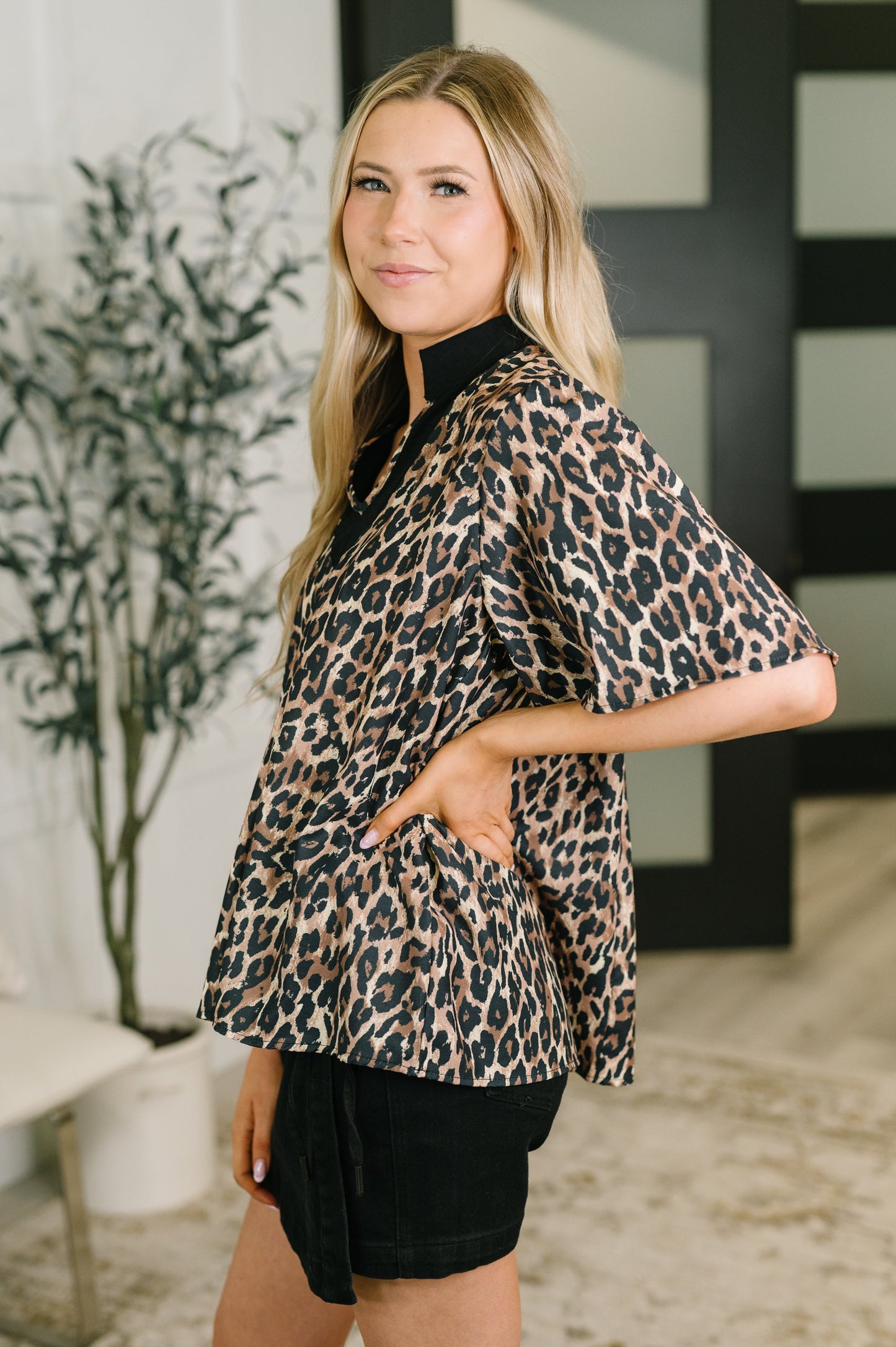 A woman modeling a leopard print short-sleeve blouse with a high black collar, paired with black shorts, standing in a modern indoor setting.