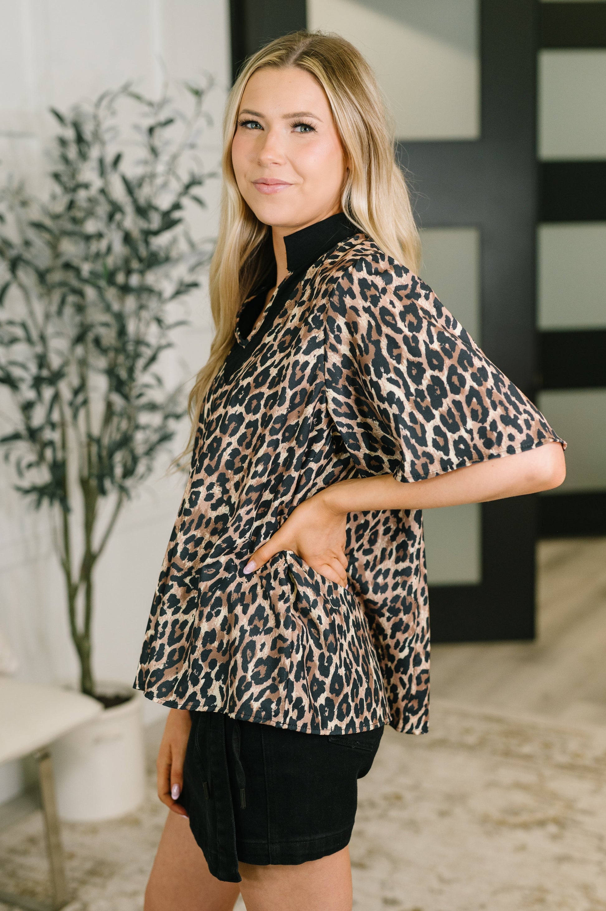 A woman modeling a leopard print short-sleeve blouse with a high black collar, paired with black shorts, standing in a modern indoor setting.
