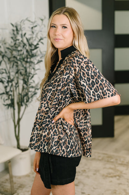A woman modeling a leopard print short-sleeve blouse with a high black collar, paired with black shorts, standing in a modern indoor setting.