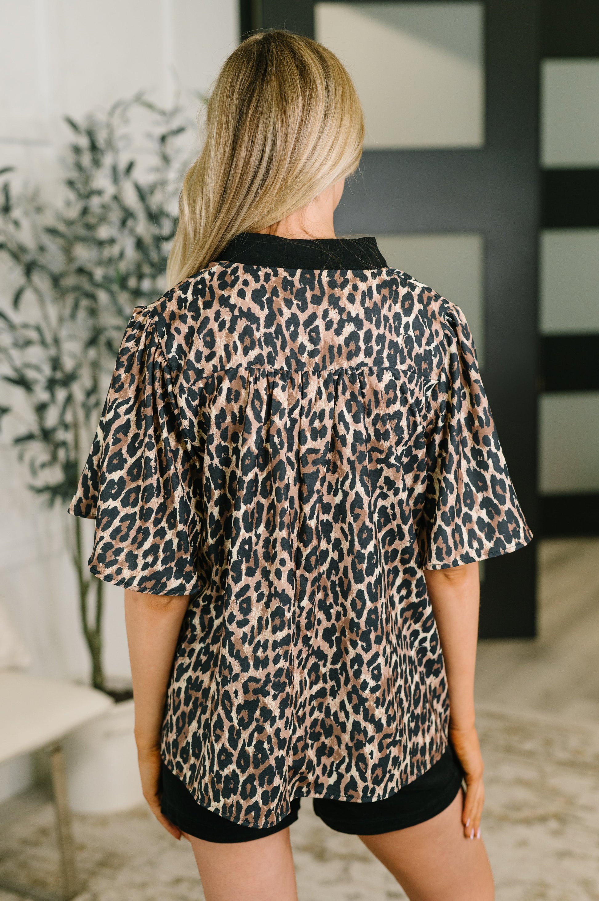 A woman shown from behind wearing a leopard print short-sleeve top with black trim, paired with black shorts, in an indoor setting.