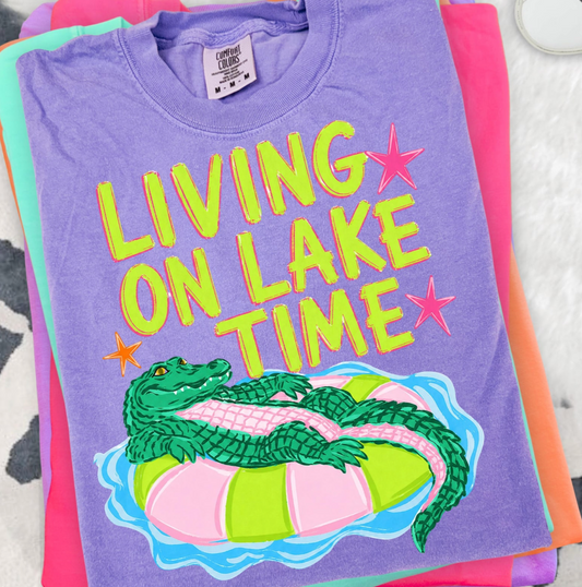 Purple t-shirt with a graphic of a alligator on an inner tube and text 'Living on Lake Time' on a colorful background.