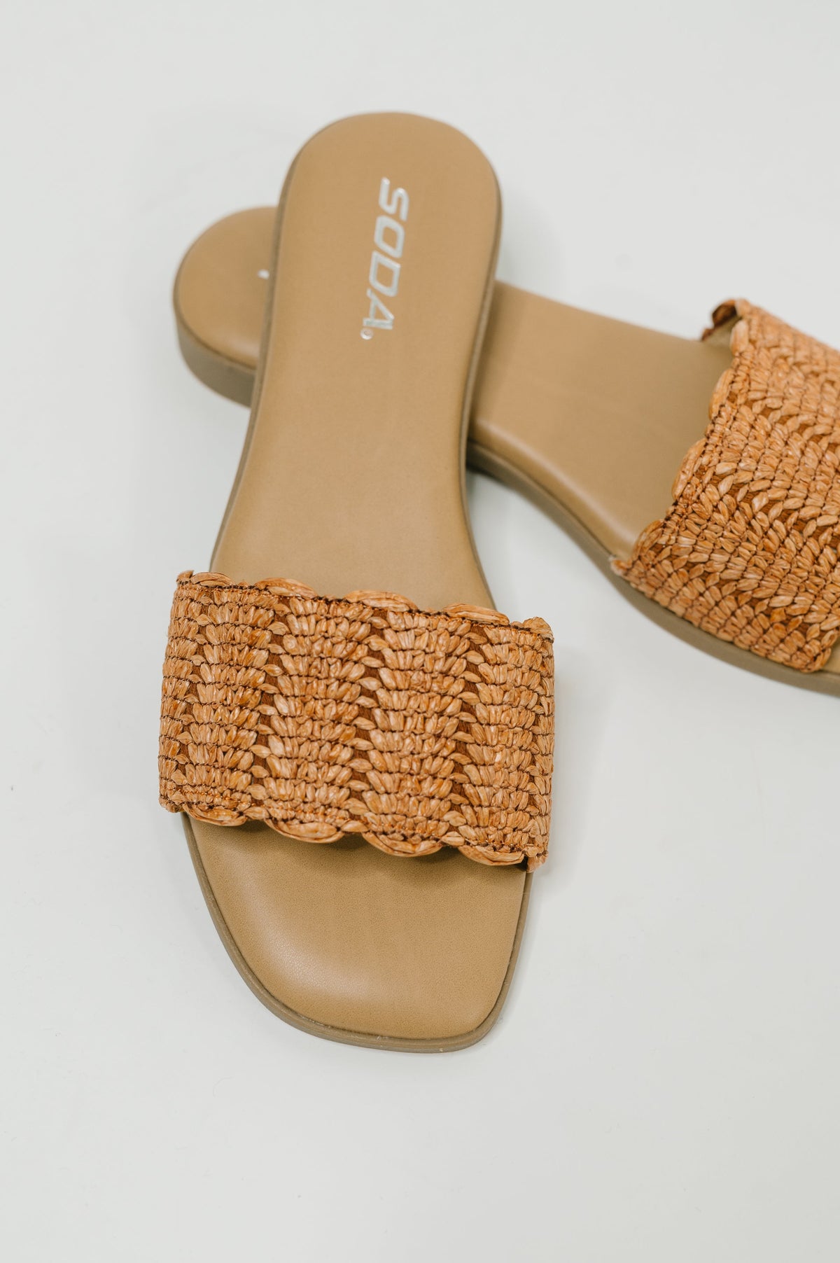 A pair of tan flat slide sandals with wide woven straps, shown on a light gray background. The brand name 'SODA.