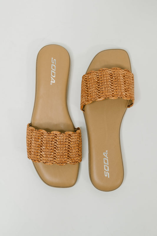 A pair of SODA slide sandals with tan woven straps and tan footbeds, shown from a top-down view on a light gray background.