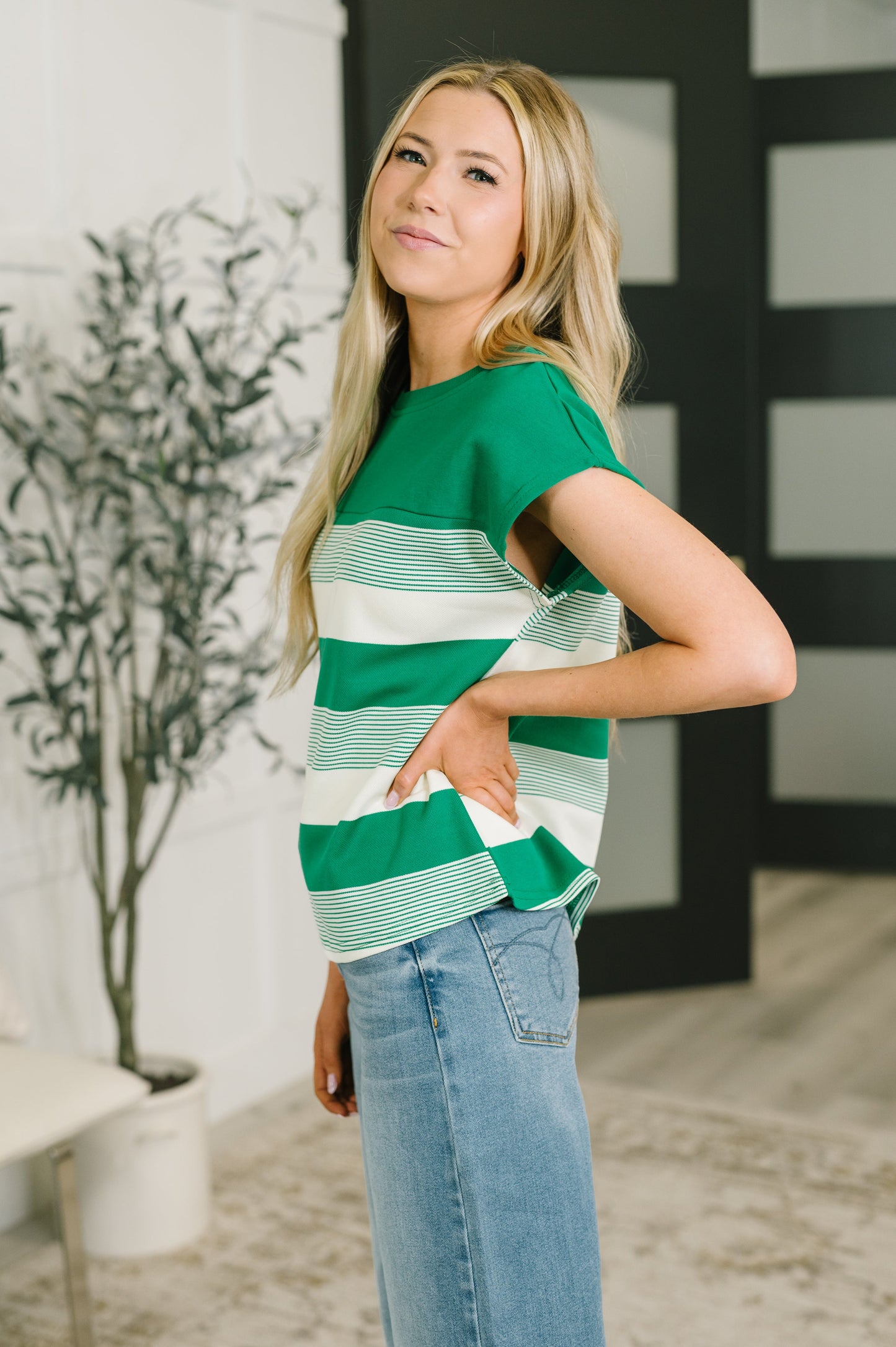A woman wearing a green and white striped short-sleeve top with a ruffled hem, paired with light blue jeans, standing in a brightly lit indoor setting.