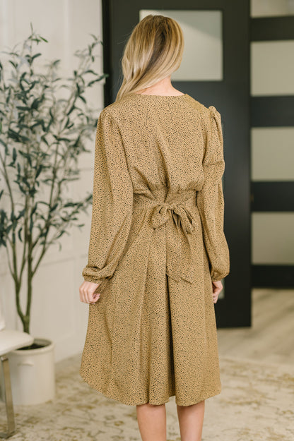 Woman wearing a brown patterned dress in an indoor setting with a plant and wall decor.