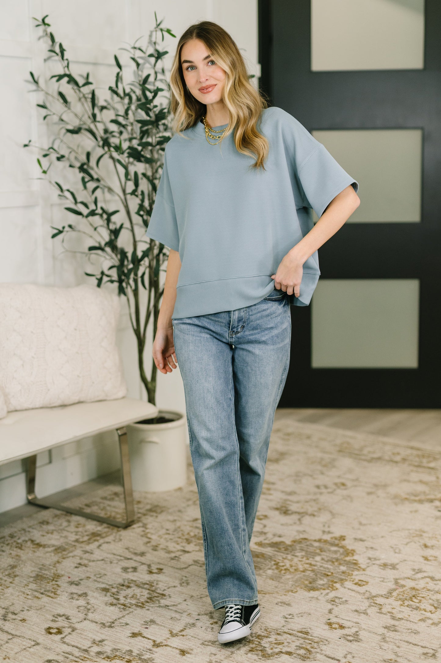Woman wearing a light blue sweater and jeans standing in a room with a plant and sofa.