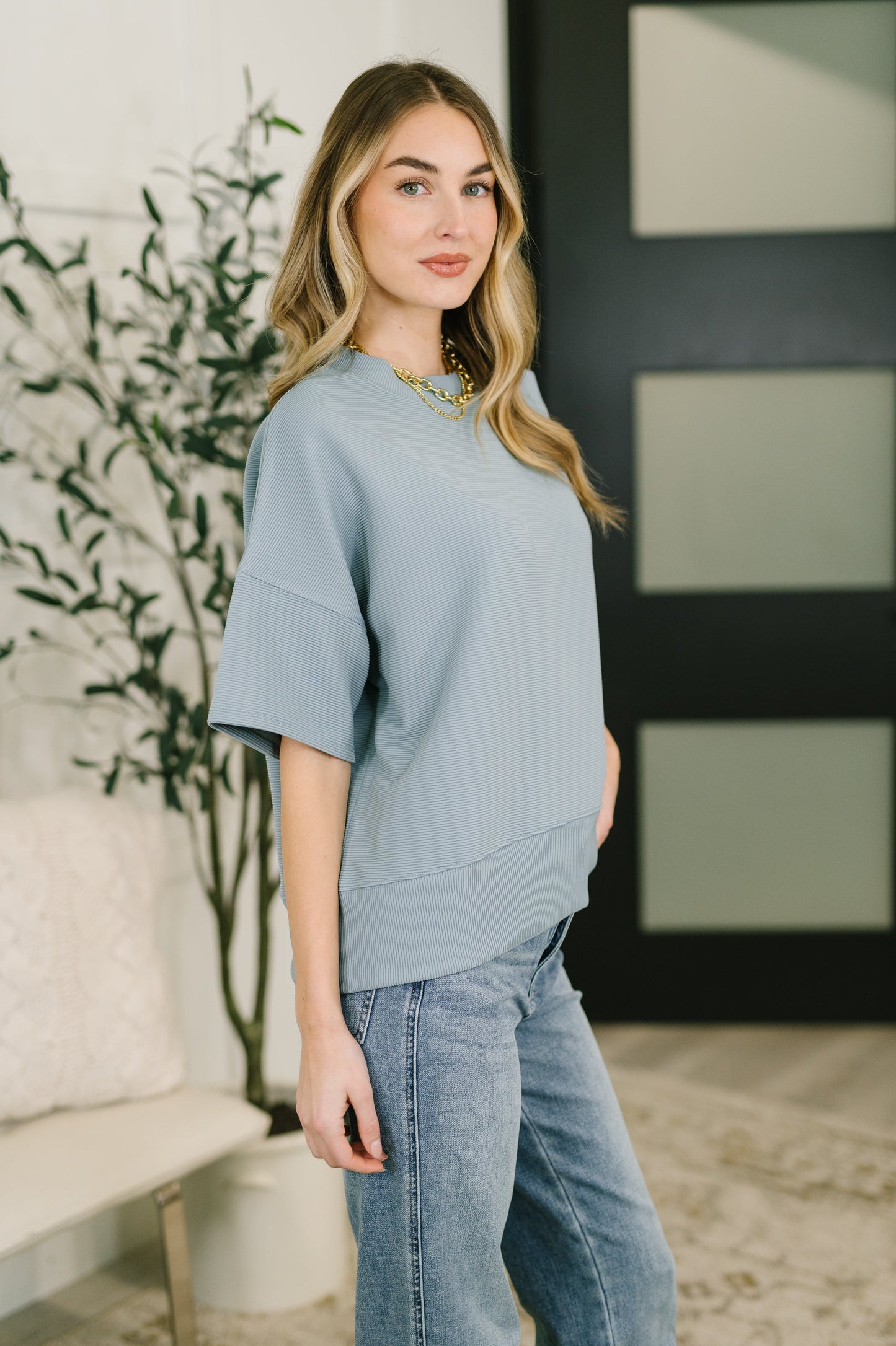 Woman wearing a light blue sweater and jeans standing indoors with a plant and wall decor in the background.
