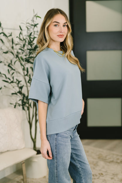 Woman wearing a light blue sweater and jeans standing indoors with a plant and wall decor in the background.