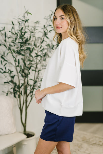 Woman wearing a white top and blue shorts standing indoors with a plant in the background