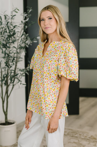 Woman wearing a floral blouse and white pants standing indoors with a plant in the background