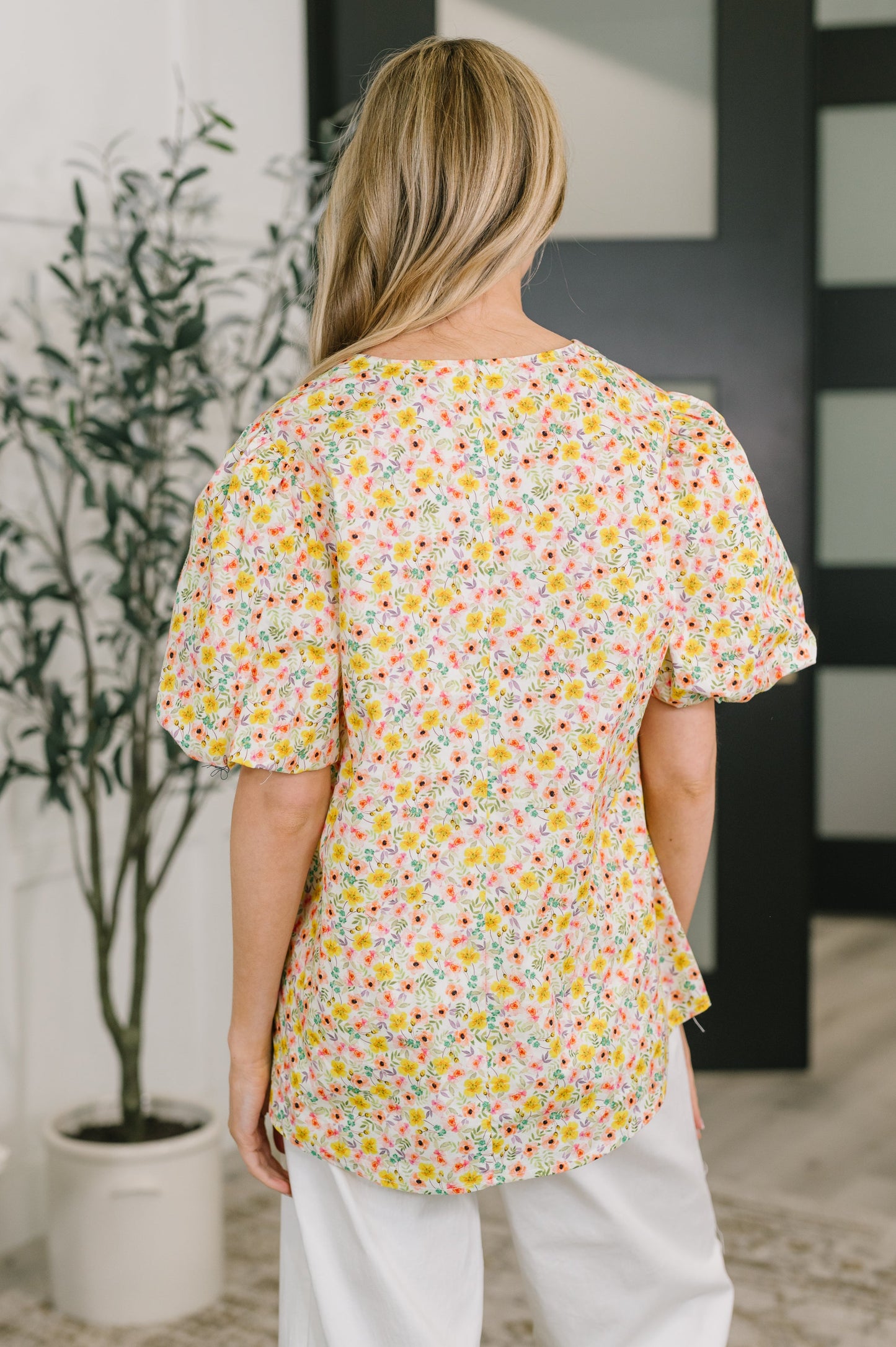 Person wearing a colorful floral blouse in an indoor setting with a plant and wall art.