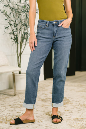 A person wearing medium-wash blue cropped jeans with rolled cuffs, paired with a mustard top and black slide sandals, standing indoors on a patterned rug.