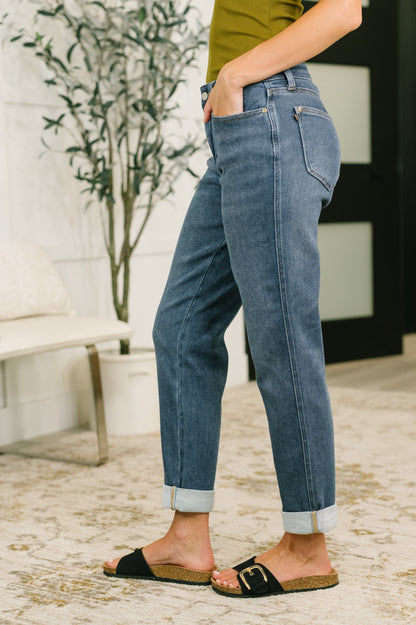 A person wearing medium-wash blue jeans with rolled cuffs, paired with black slide sandals and an olive green top, standing in a modern indoor setting with a patterned rug and a potted plant.