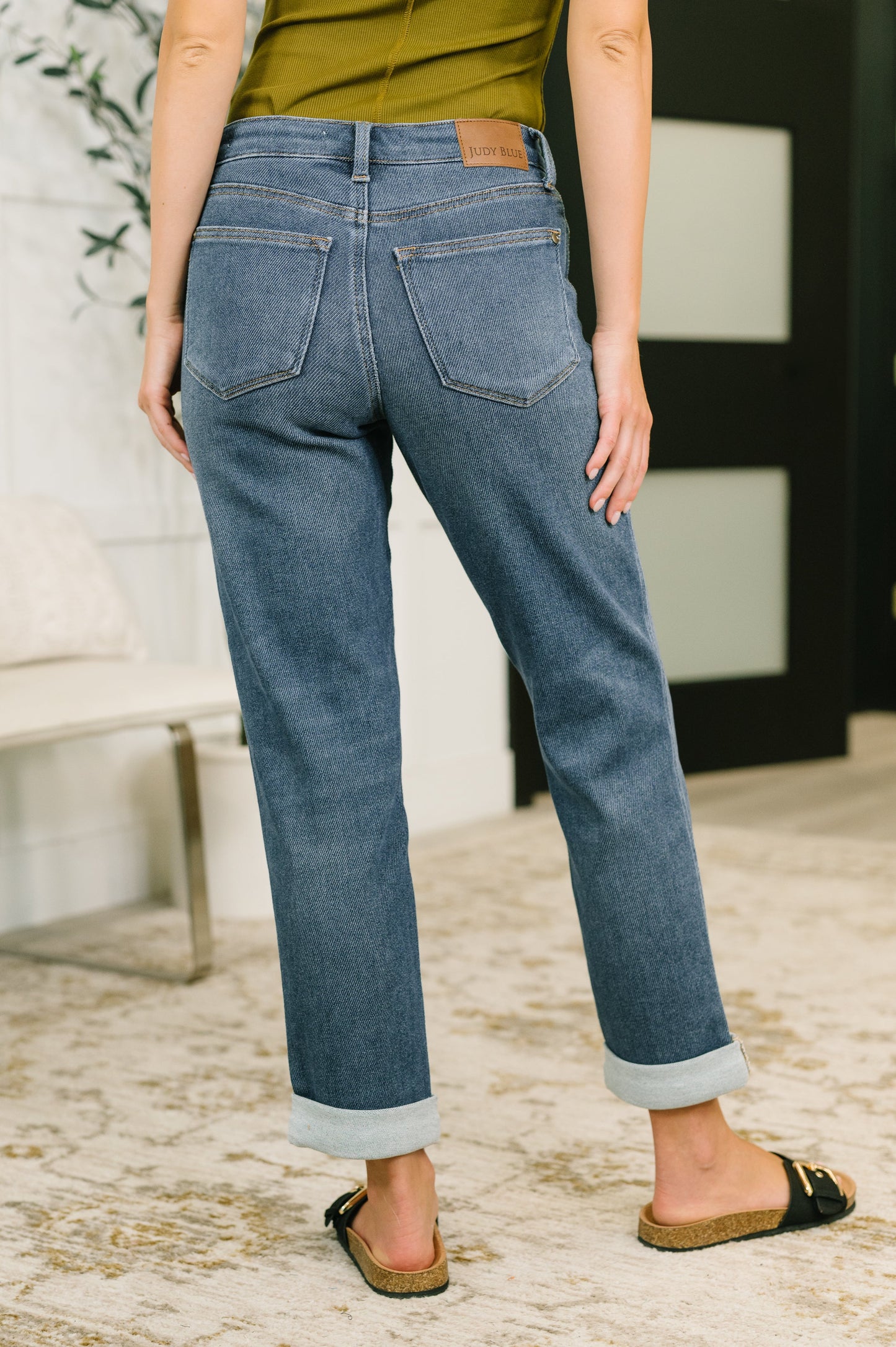 A person shown from the back wearing medium-wash blue cuffed jeans, an olive green top, and black slide sandals in an indoor room setting.
