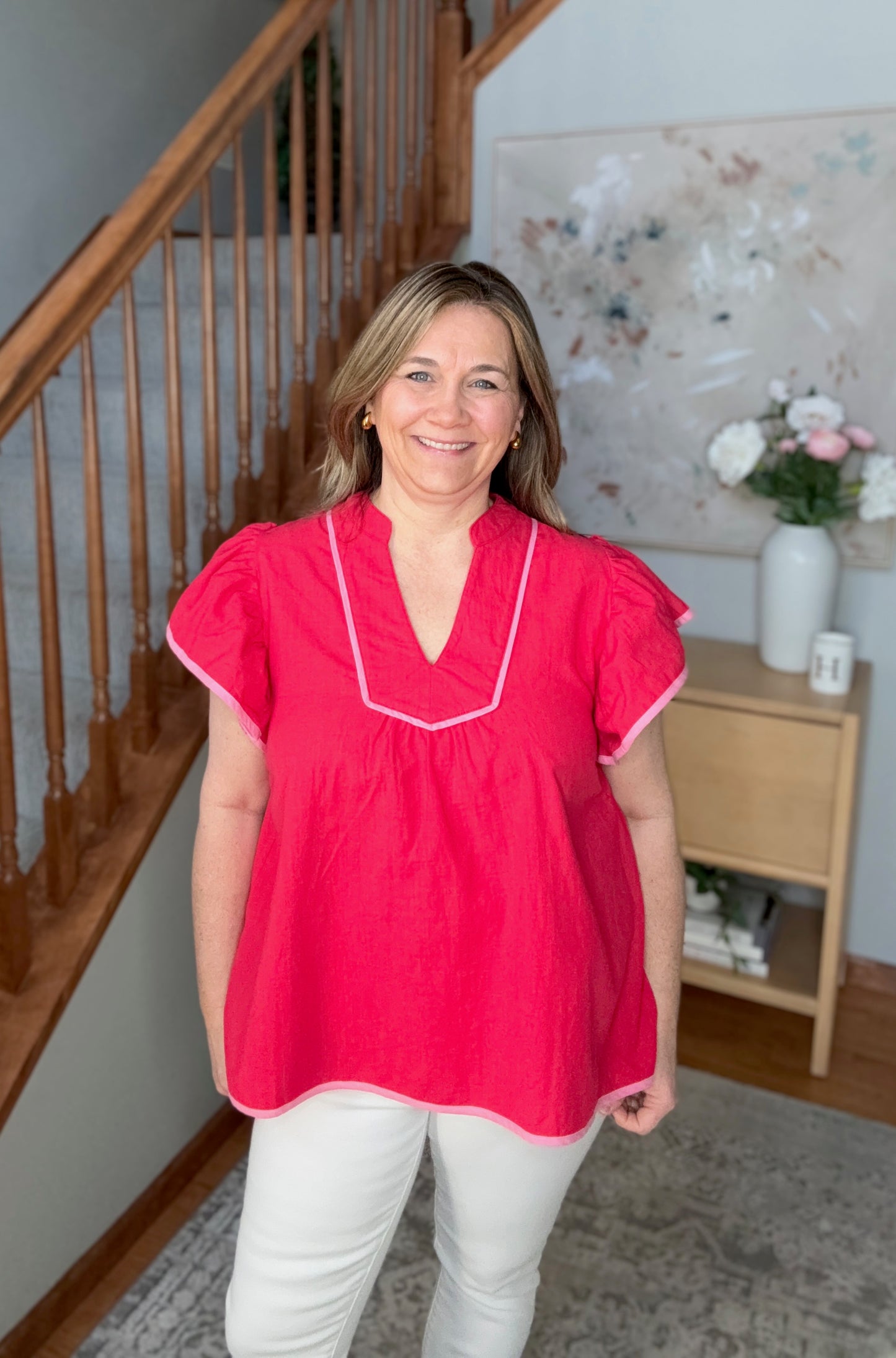 Woman wearing a red blouse standing in a home setting with a staircase and decor in the background.