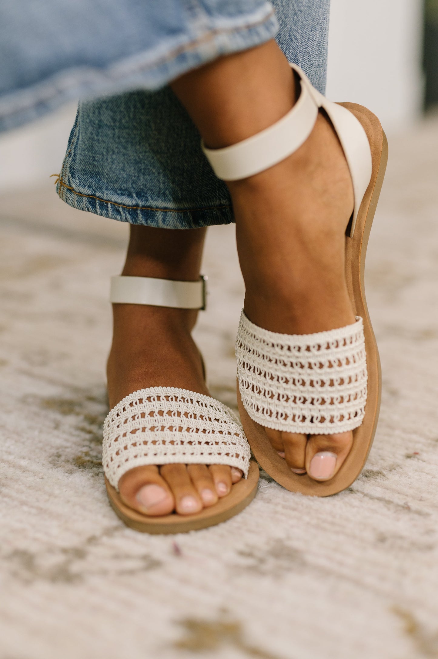 Brown sandals with white embellishments worn with blue jeans on a stone surface.