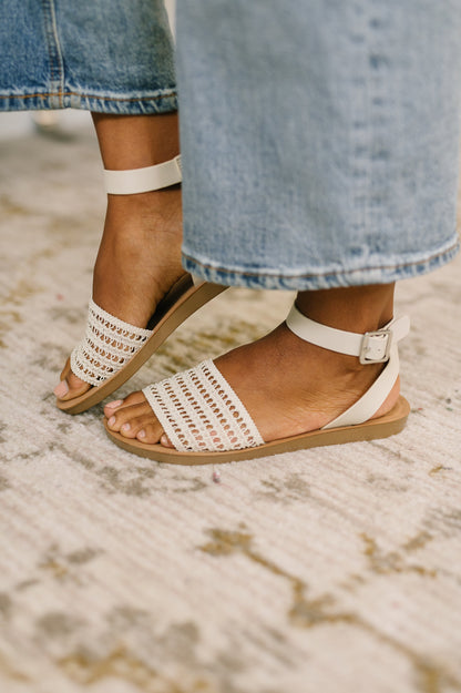 White sandals with brown soles worn with rolled-up jeans on a textured floor.